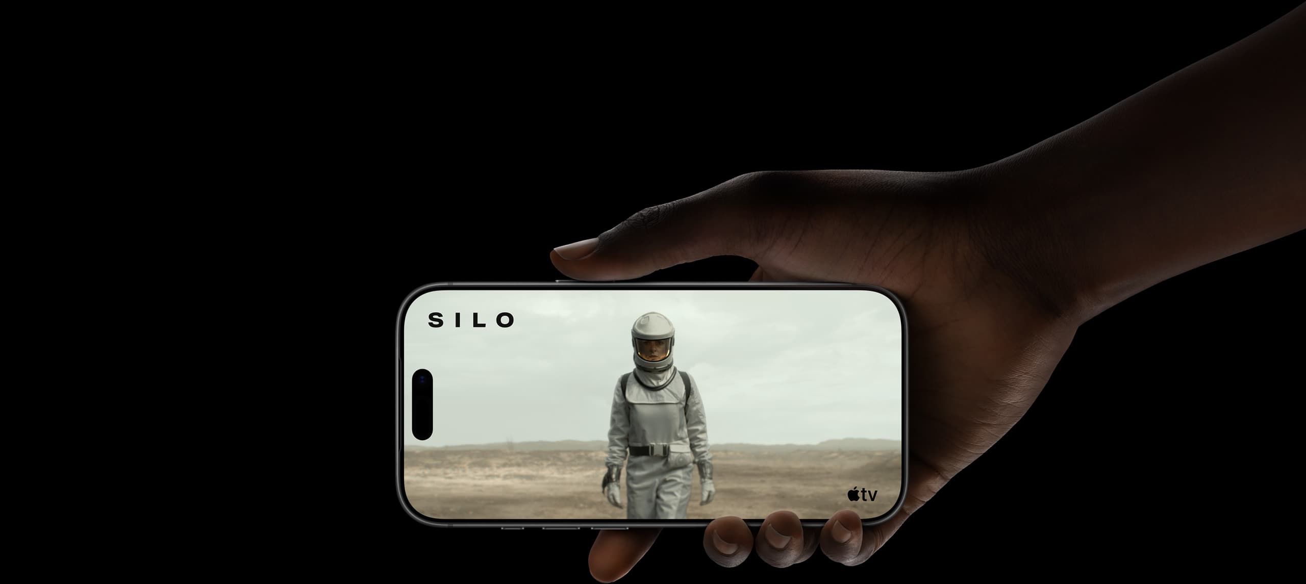 A hand holding iPhone 17 Pro, displaying a woman wearing a protective suit and helmet, walking through a barren landscape. Apple TV logo, series title: Silo